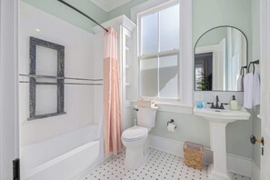 Un baño con inodoro y lavamanos blancos, ducha con cortina rosa, espejo y ventana.