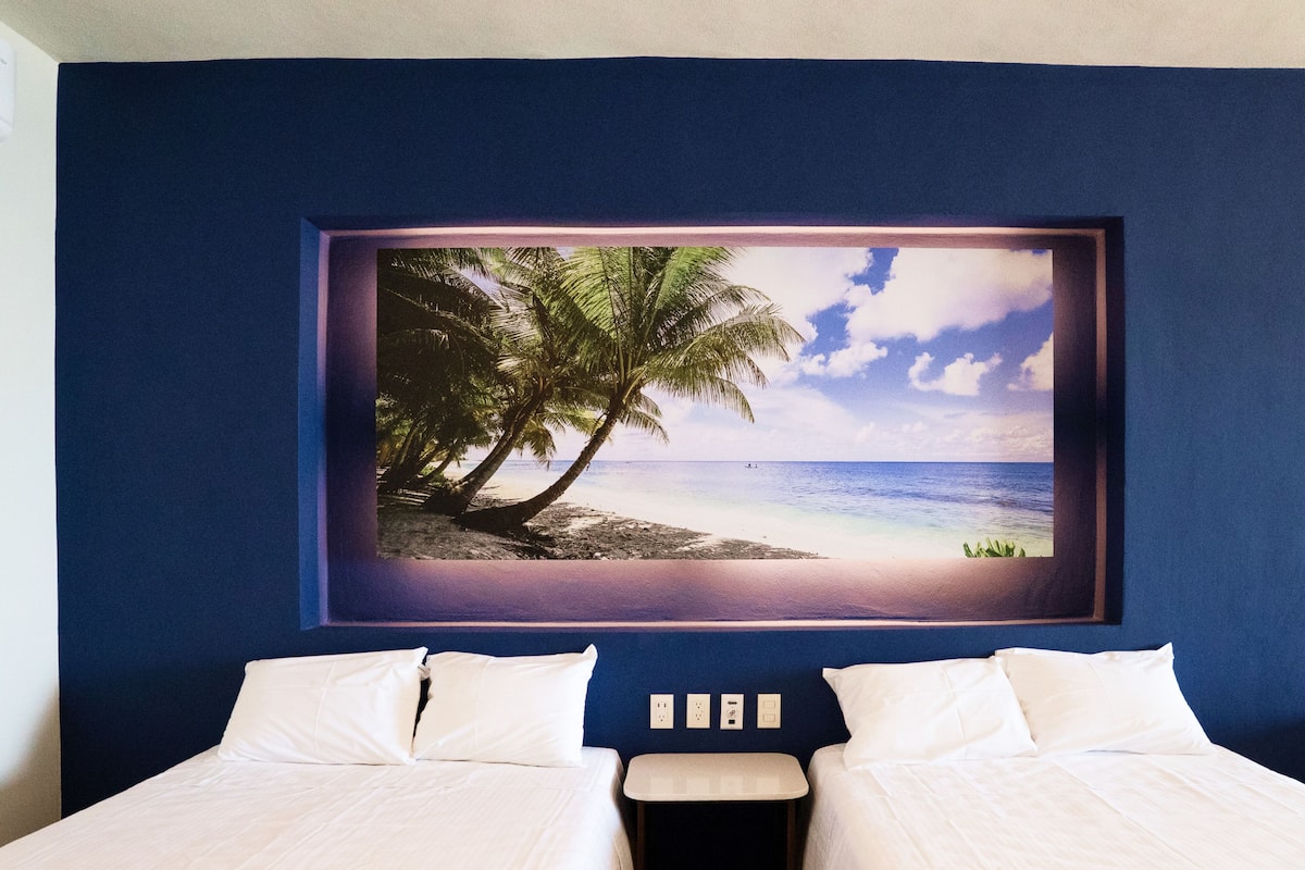 Un dormitorio con un cuadro grande enmarco de una playa, dos camas y una mesita de noche con una lámpara.