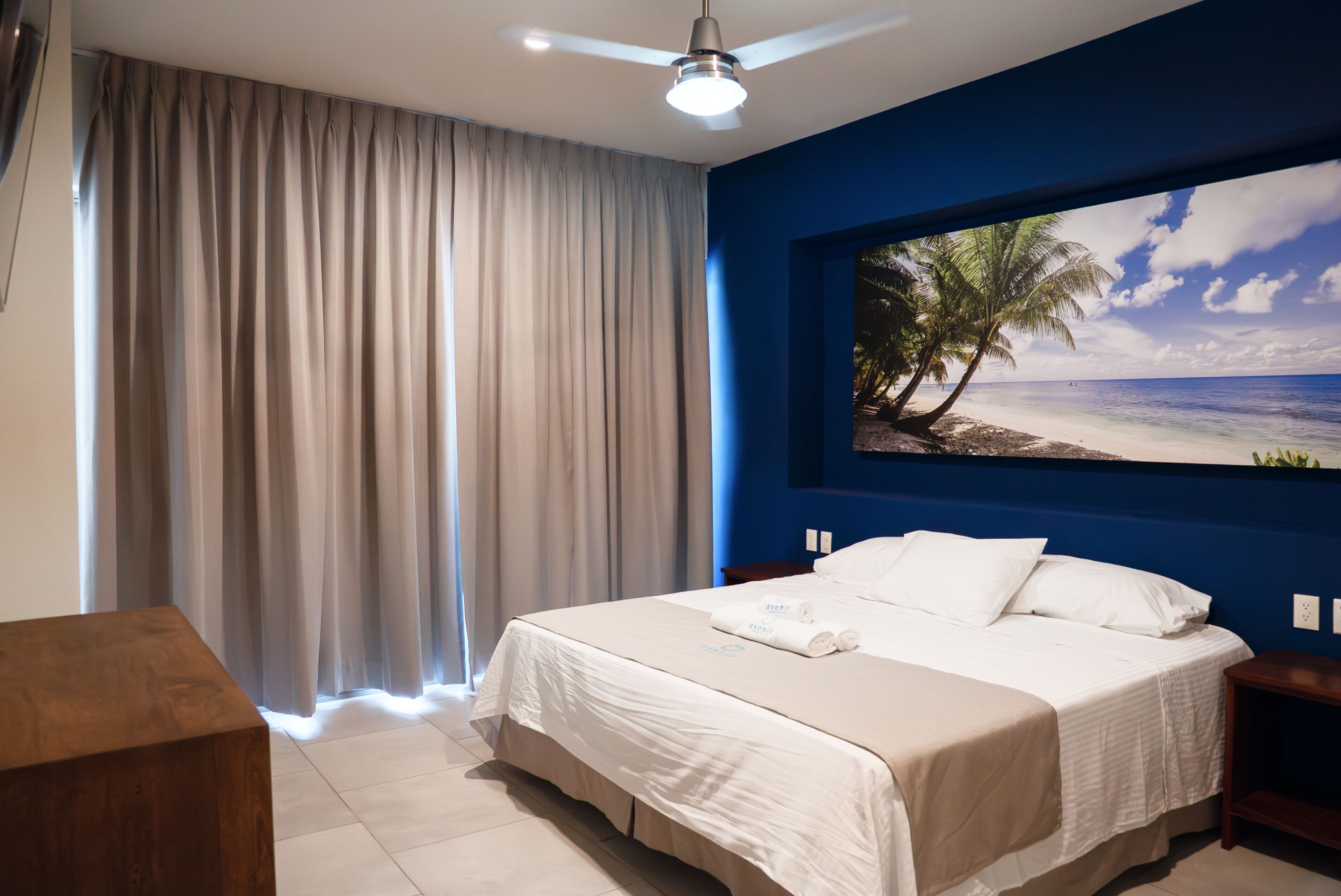Una habitación de hotel con cama, cortinas, ventilador de techo, un cuadro de una playa y una mesita de noche de madera.