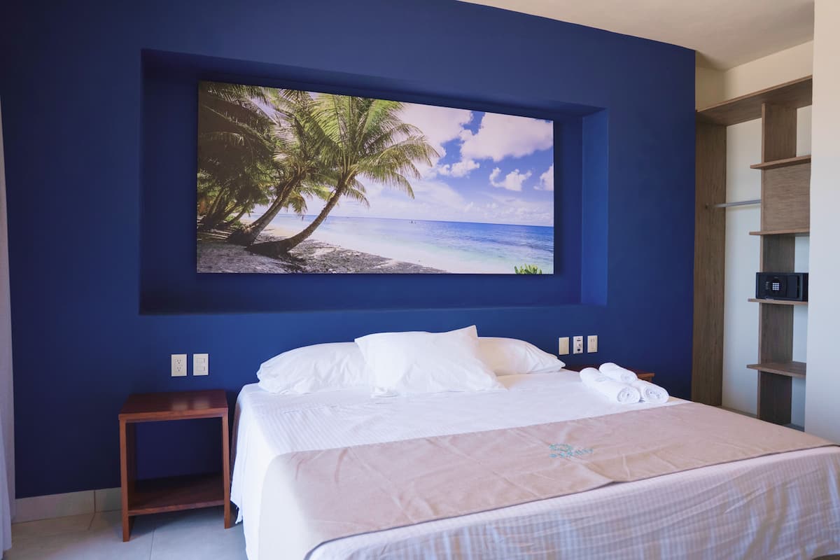 Un dormitorio con una cama grande, una mesita de noche, un televisor con pantalla montada en la pared mostrando una escena de playa y una estantería de madera.