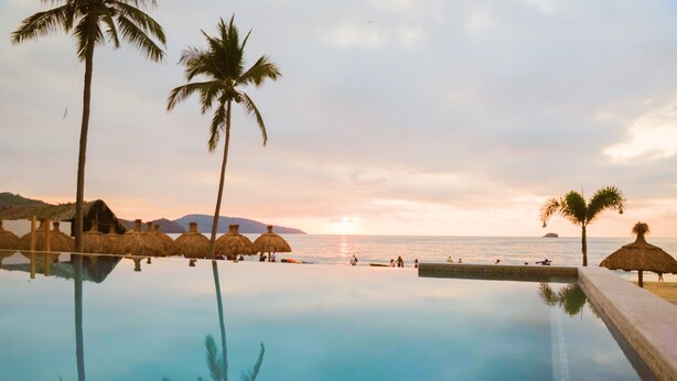 An infinity pool with palm trees and thatched umbrellas, overlooking the sea at sunset. - Avenir Armonia Suites (Rincón de Guayabitos)