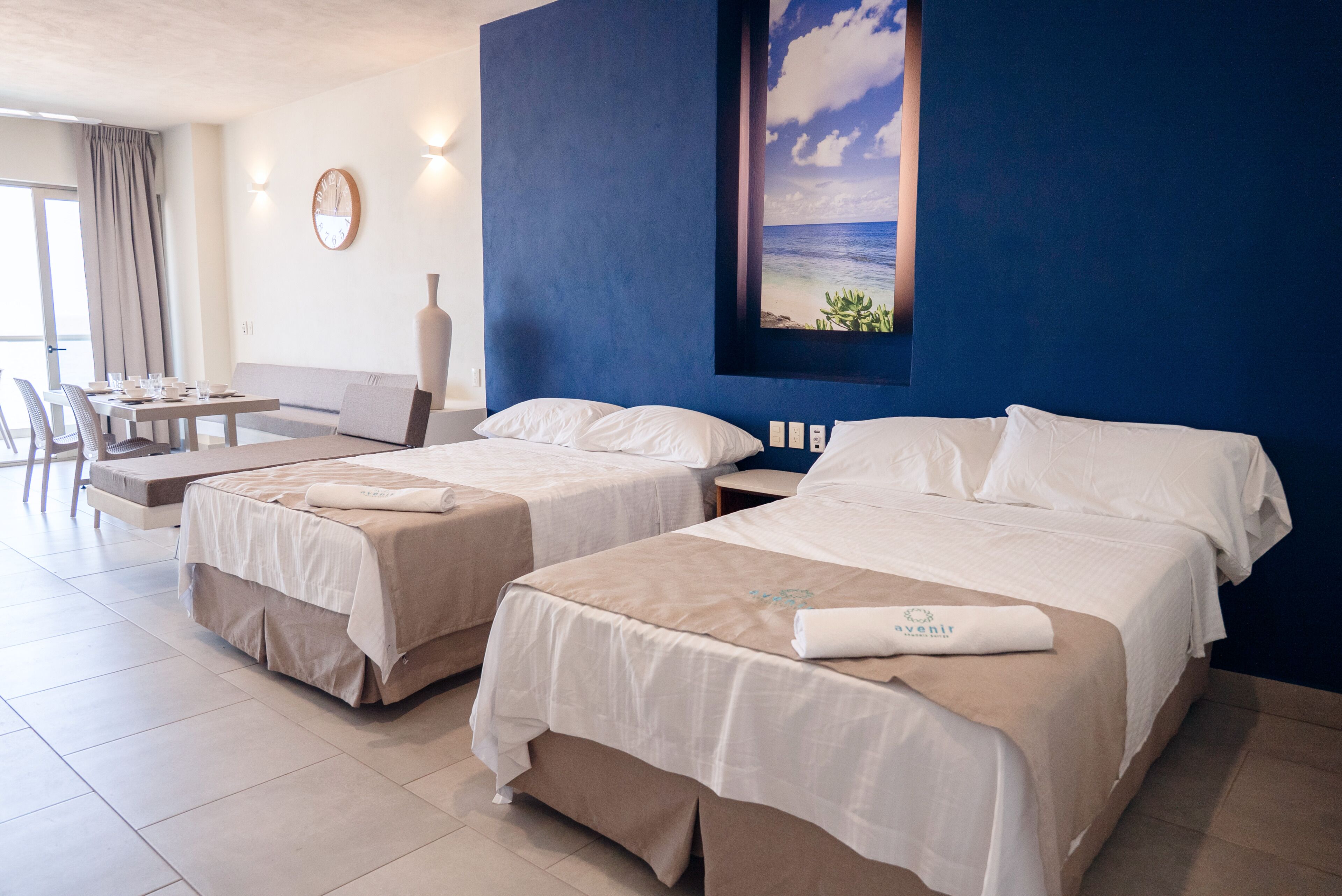 A hotel room with two beds, a dining area, and a large window with a view of the ocean.