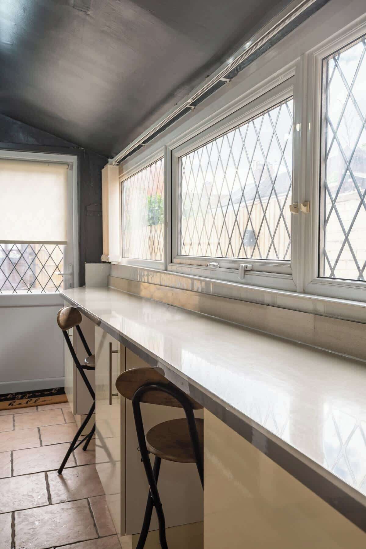 A kitchen with a white countertop, two bar stools, and a window with a diamond-patterned screen.