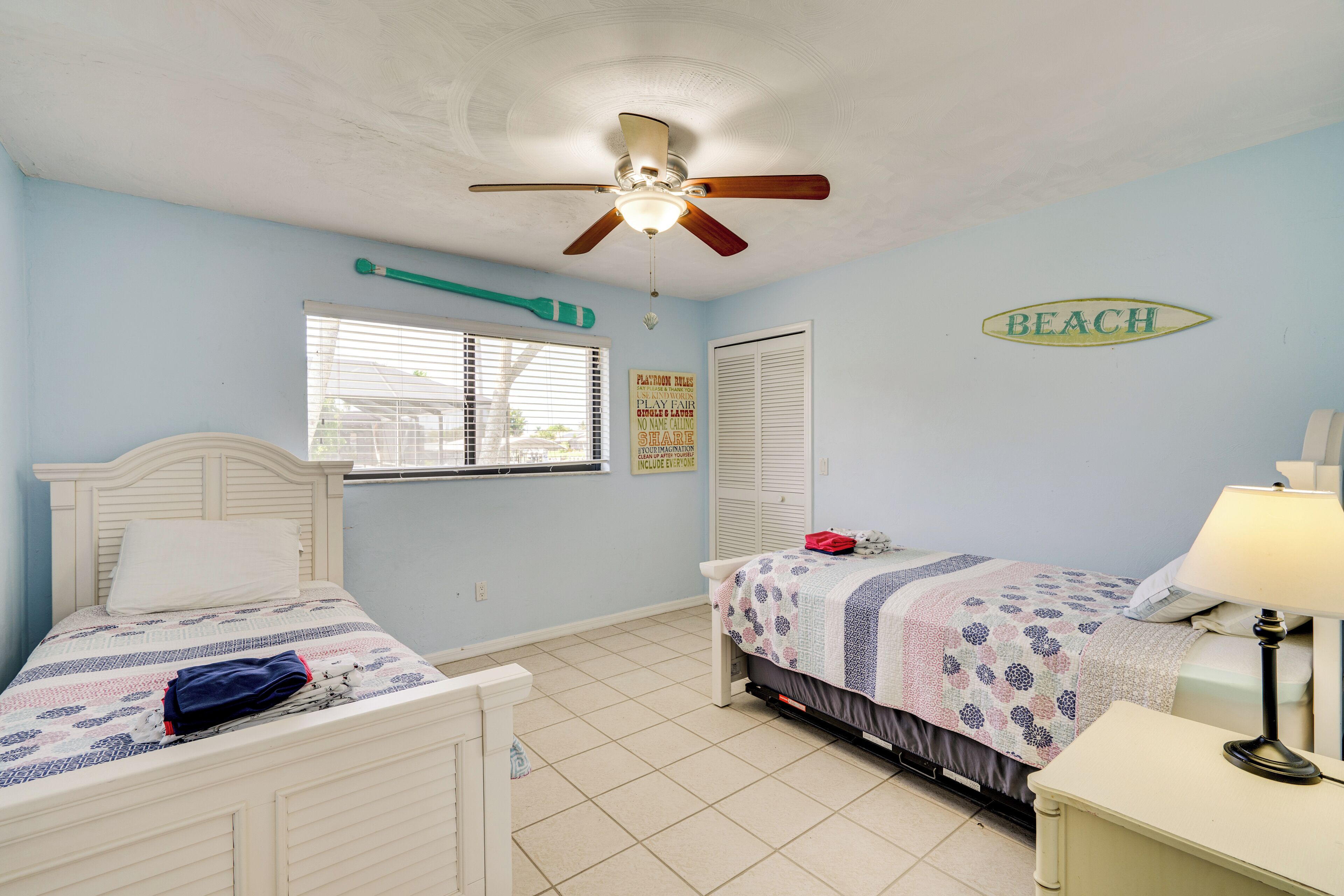 Une chambre avec deux lits, un ventilateur de plafond, une fenêtre avec des stores, un panneau indiquant « PLAGE », et une table de chevet avec une lampe.