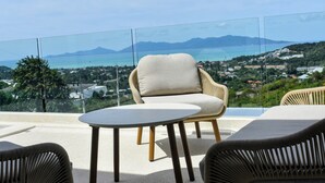 A balcony with a view of a coastal town, furnished with a chair and a small table.