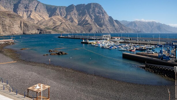 Un port de plaisance sur la côte, avec des bateaux amarrés, une rive rocheuse et des montagnes en arrière-plan.