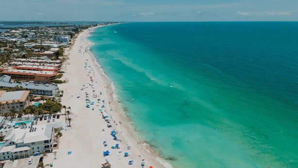 Vista aérea de una playa con una extensa franja de arena, algunos edificios y un océano azul cristalino.