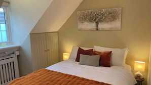 A bedroom with a sloped ceiling, a bed with white bedding and orange throw pillows, a painting of a tree on the wall, and a window with blinds. - The Frog, Langport Luxury (Huish Episcopi)
