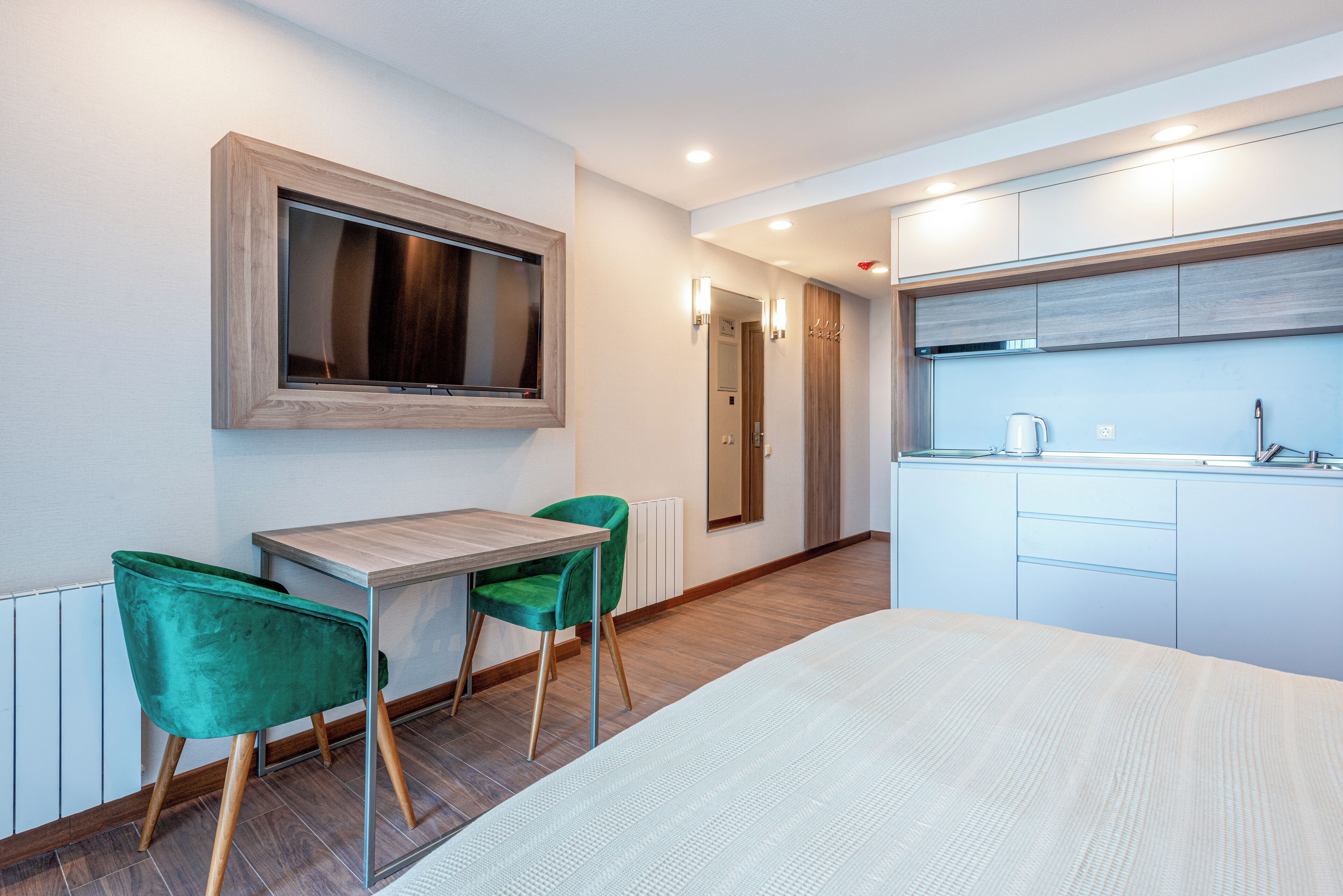 A modern hotel room with a bed, a desk with two green chairs, a TV mounted on the wall, a kitchen area with a kettle, and a radiator.