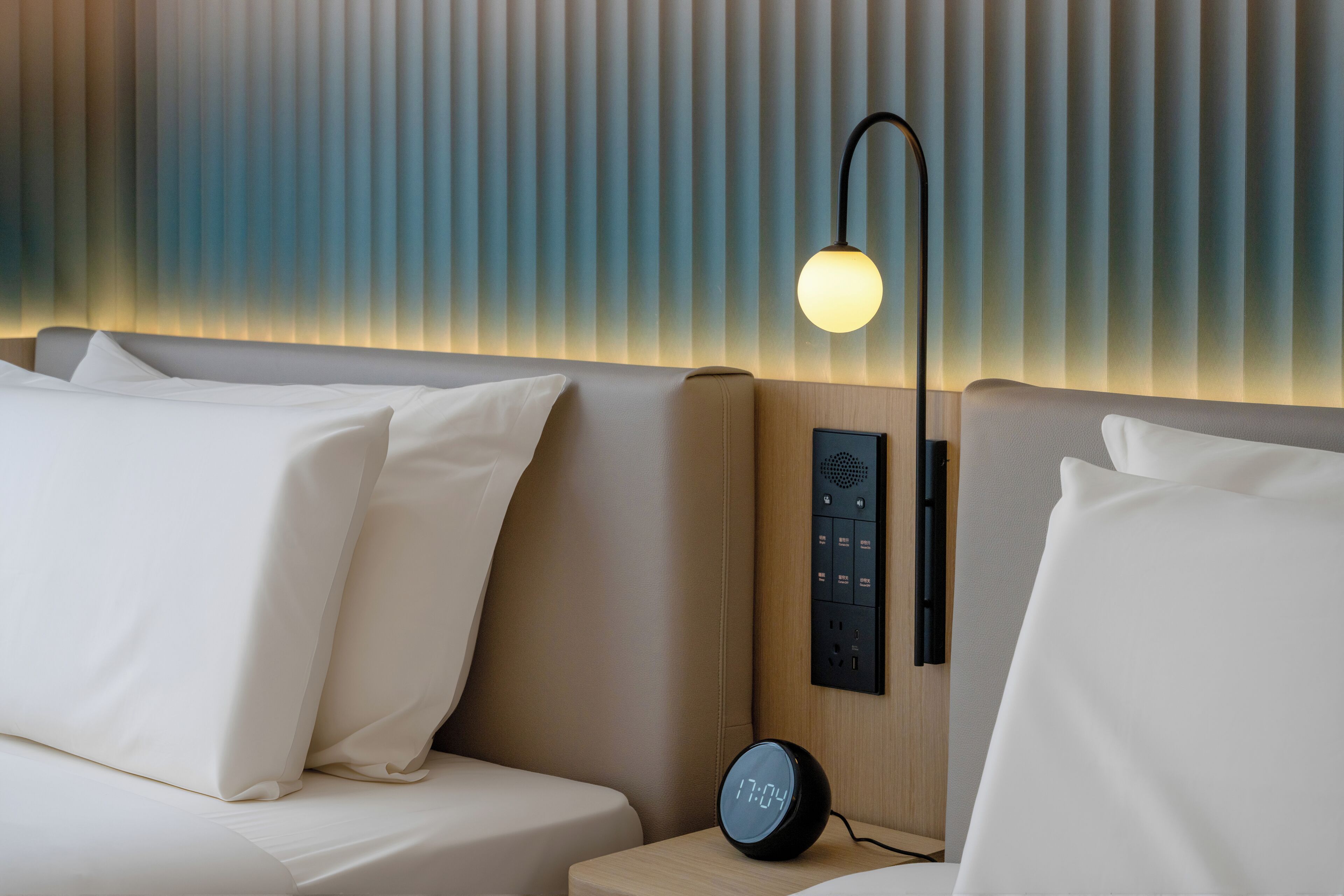 A modern hotel room with a bed, a bedside table with a clock, a wall-mounted control panel, and a wall-mounted lamp.