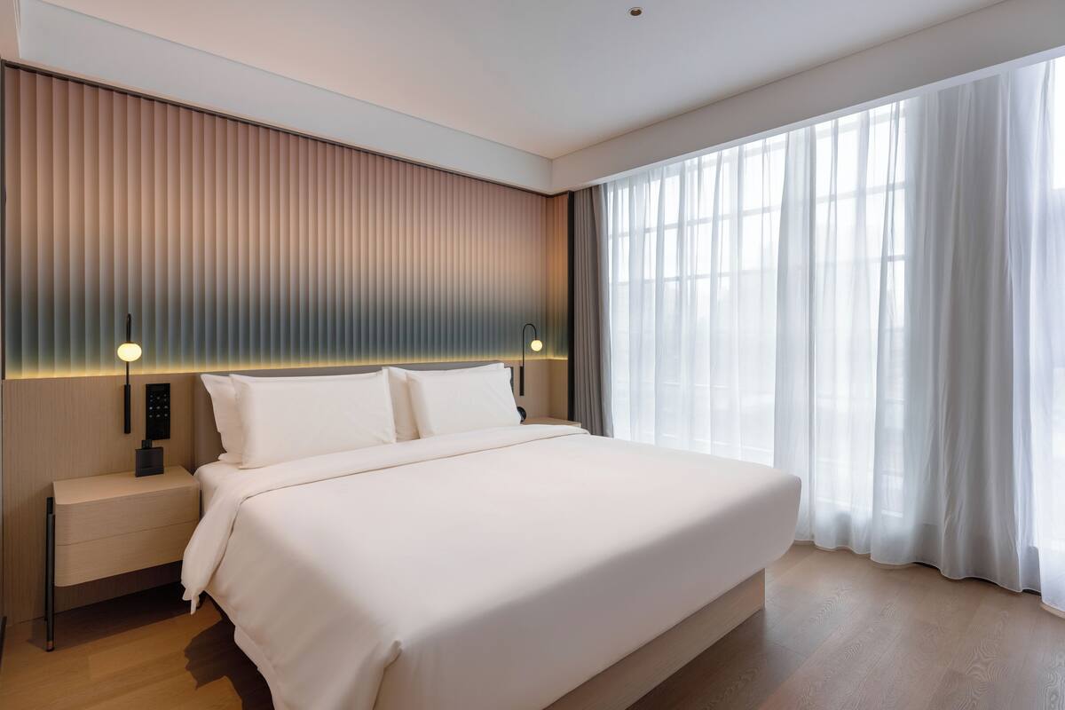 Ein modernes Hotelzimmer mit einem großen Bett, einem Nachttisch mit Lampe und Blick ins Freie durch transparente Vorhänge.