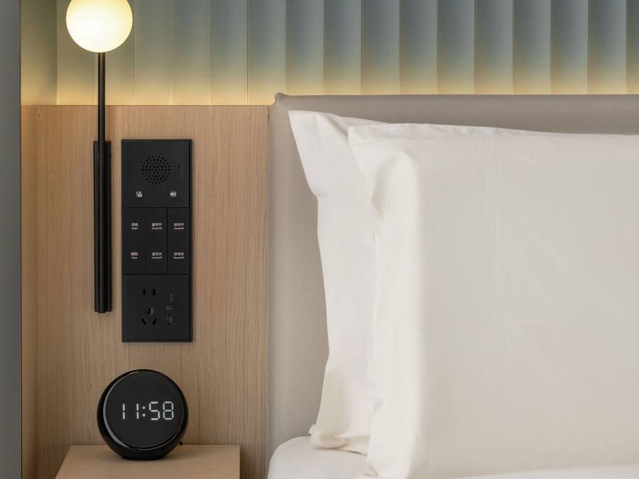 A modern hotel room with a bed, a bedside table with a digital clock, a lamp, and a control panel.