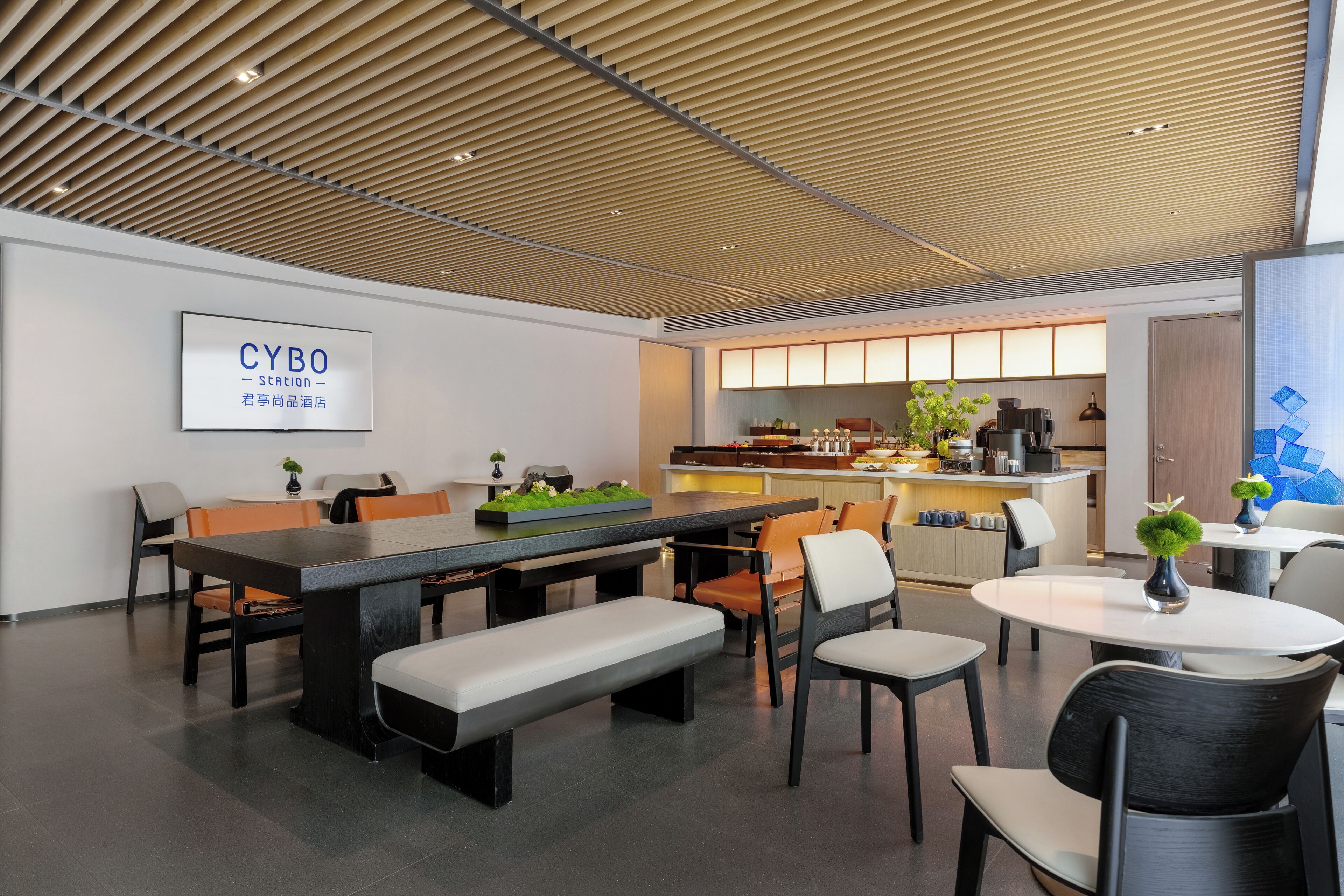 A modern hotel lobby with a wooden slatted ceiling, a reception desk, a dining area with a long table and chairs, and a kitchen area in the background.