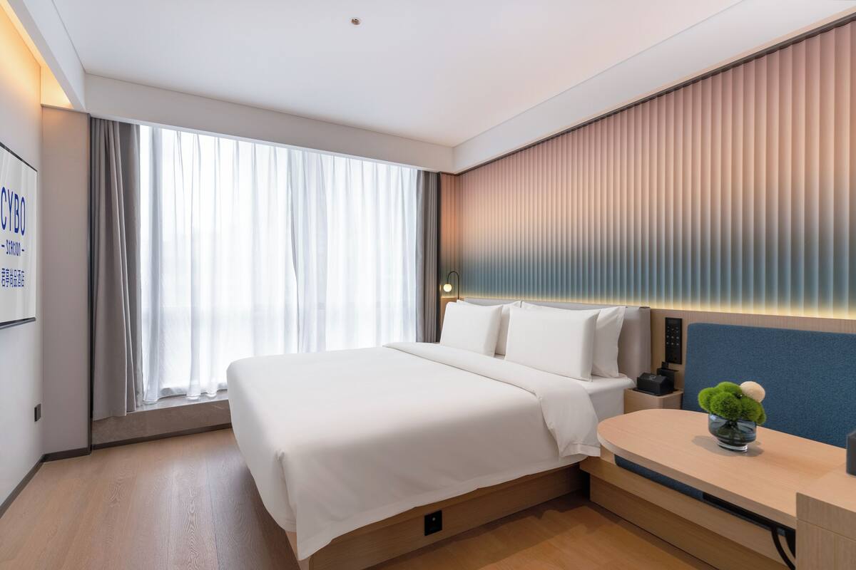 Ein modernes Hotelzimmer mit einem großen Bett, einem Nachttisch mit Lampe und Blick auf die Stadt durch große Fenster.