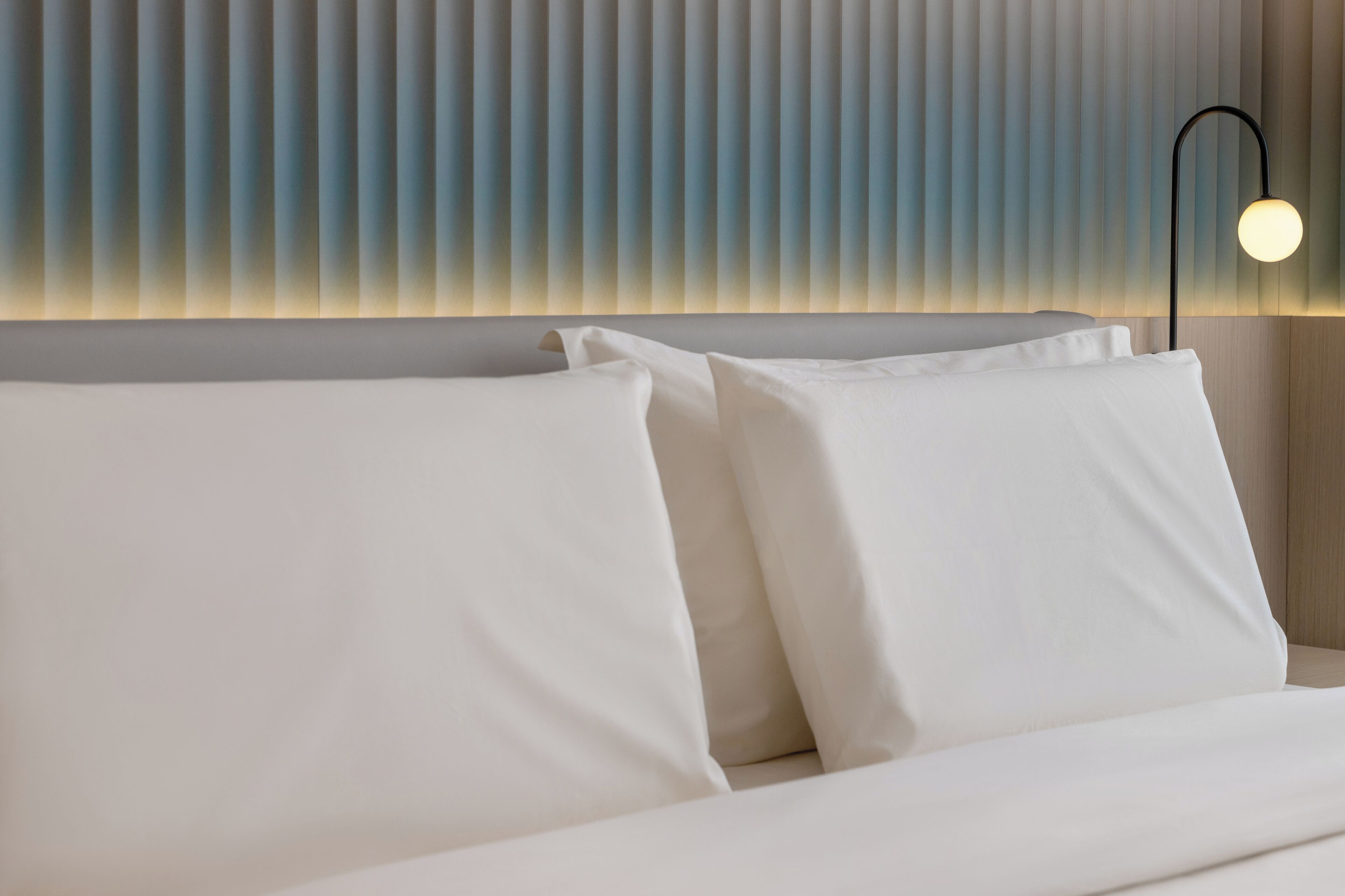 A neatly made bed with white linens, a wall with vertical blinds, and a modern floor lamp.
