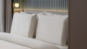Un lit bien fait, avec du linge de lit blanc, une lampe de chevet et des stores verticaux à la fenêtre.