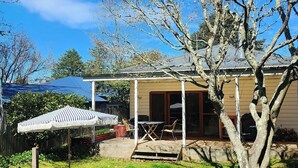 Ein überdachter Patio-Bereich mit Tisch und Stühlen, einem gestreiften Sonnenschirm und einem Baum im Vordergrund.