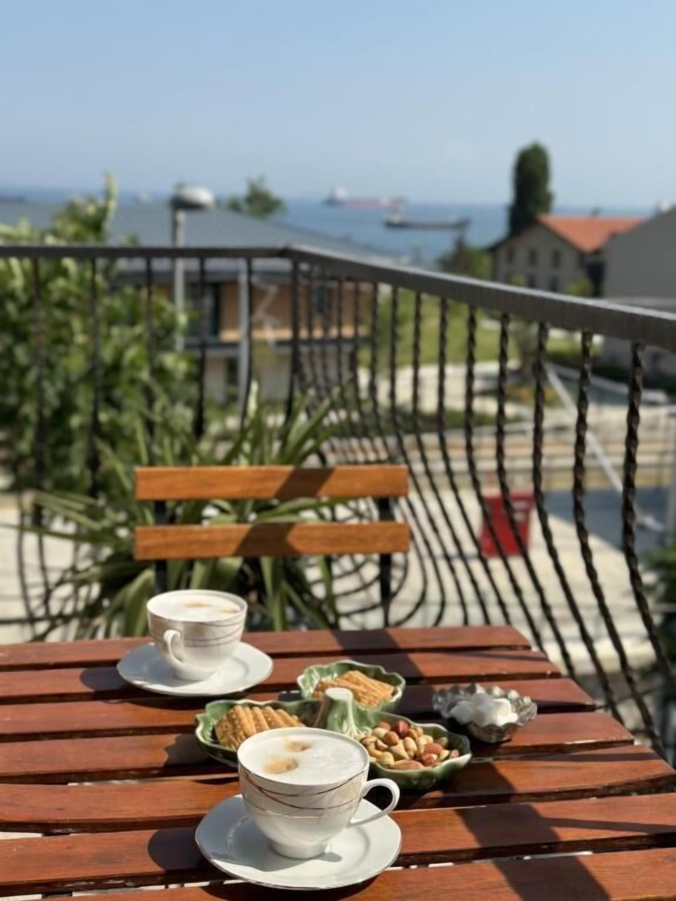 Ein Holztisch mit zwei Kaffeetassen und einer Schüssel mit Snacks auf einem Balkon mit Blick auf ein Gewässer und Gebäude.