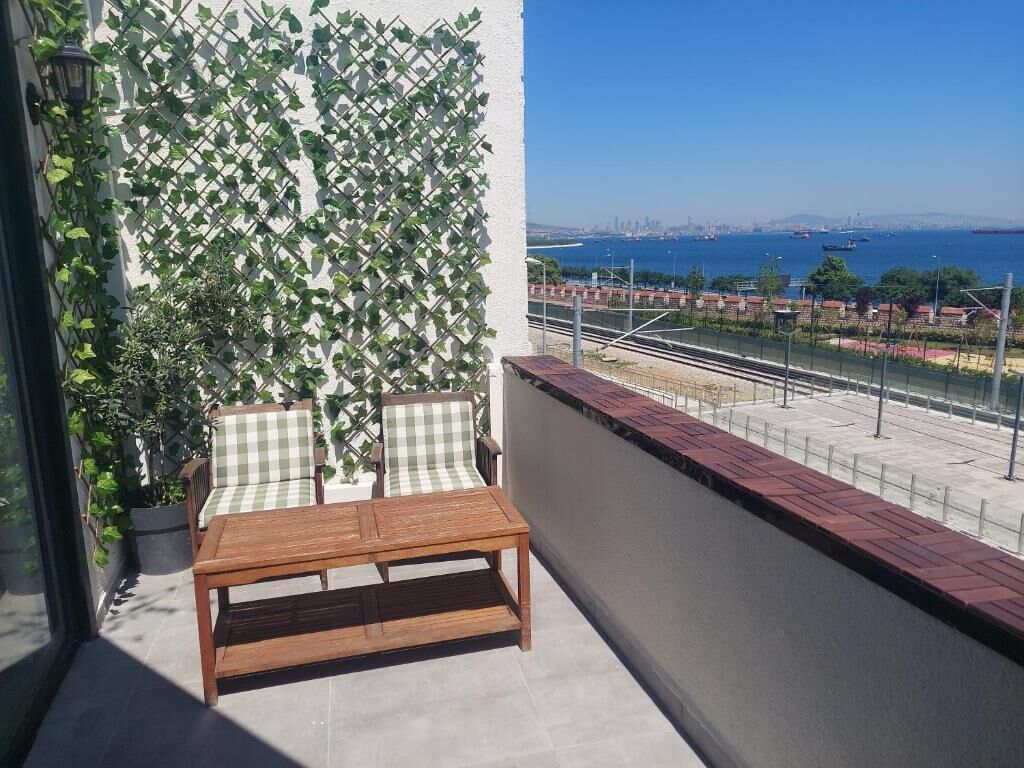 Un balcón con mesa de madera y dos sillas, un muro ajedrezado con plantas trepadoras, y vistas a un paisaje urbano y un cuerpo de agua.