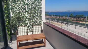 Un balcón con mesa de madera y dos sillas, un muro ajedrezado con plantas trepadoras, y vistas a un paisaje urbano y un cuerpo de agua.
