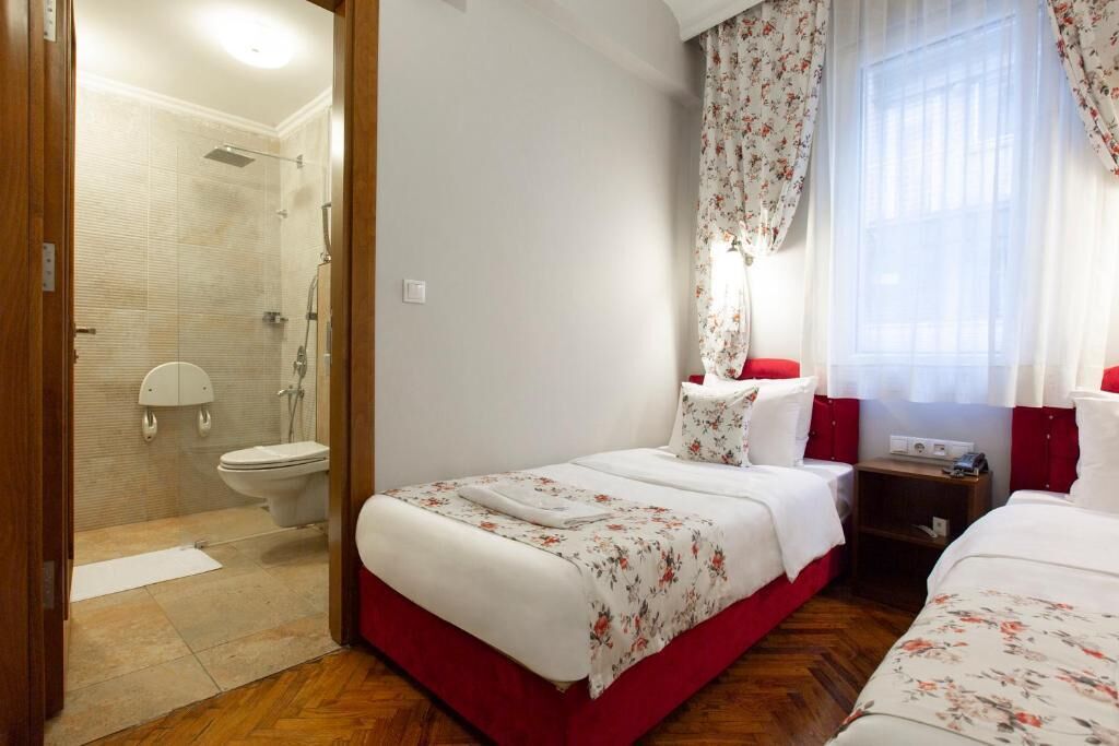 Quarto de hotel com área de chuveiro, cama com roupa de cama floral, mesa de cabeceira e janela com cortinas.