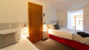 Une chambre d’hôtel avec deux lits, une porte en bois, un interrupteur et une vue sur le couloir.