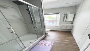 A bathroom with a glass shower enclosure, a toilet, a sink, and a window with a view of a building outside.