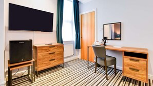 Une chambre d’hôtel équipée d’un téléviseur à écran plat, d’un bureau en bois, d’une chaise, d’une commode, d’un miroir et d’une fenêtre avec des rideaux.