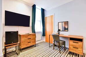 Ein Hotelzimmer mit Flachbildfernseher, Schreibtisch aus Holz, Stuhl, Kommode, Spiegel und einem Fenster mit Vorhängen.