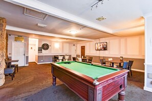 Een biljart in een ruimte met vloerbedekking, stoelen, een dartbord en een aan de muur bevestigde televisie.