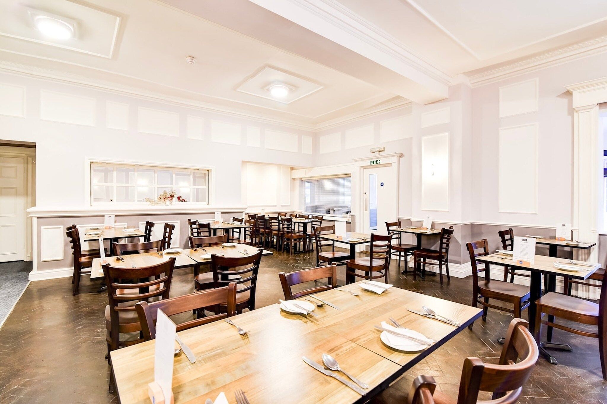 A spacious dining area with multiple tables and chairs, a white interior, and a large window providing natural light.
