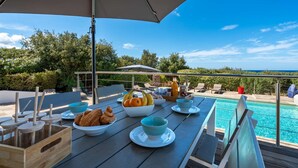 A dining area with a pool view, featuring a table with breakfast items, a large umbrella, and outdoor seating. - Villa Castille – Pool and Sea View in Lumio, Balagne (LUMIO)