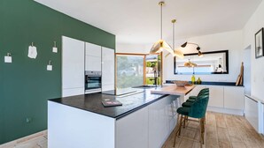 A modern kitchen with a central island, a dining area with a wooden table, and a large window offering a view of the outdoors. - Villa Castille – Pool and Sea View in Lumio, Balagne (LUMIO)