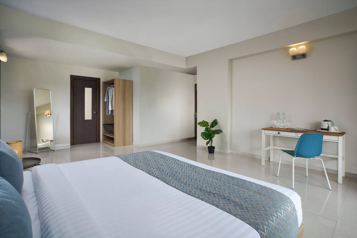 Habitación de hotel con una cama grande, un escritorio con silla, un espejo, un armario y una planta en maceta.