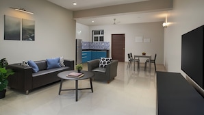 A modern living room with a sofa, coffee table, and a TV. There is a dining area with chairs and a table. The kitchen is visible in the background with blue cabinets. - Paprika Stays (Panaji)