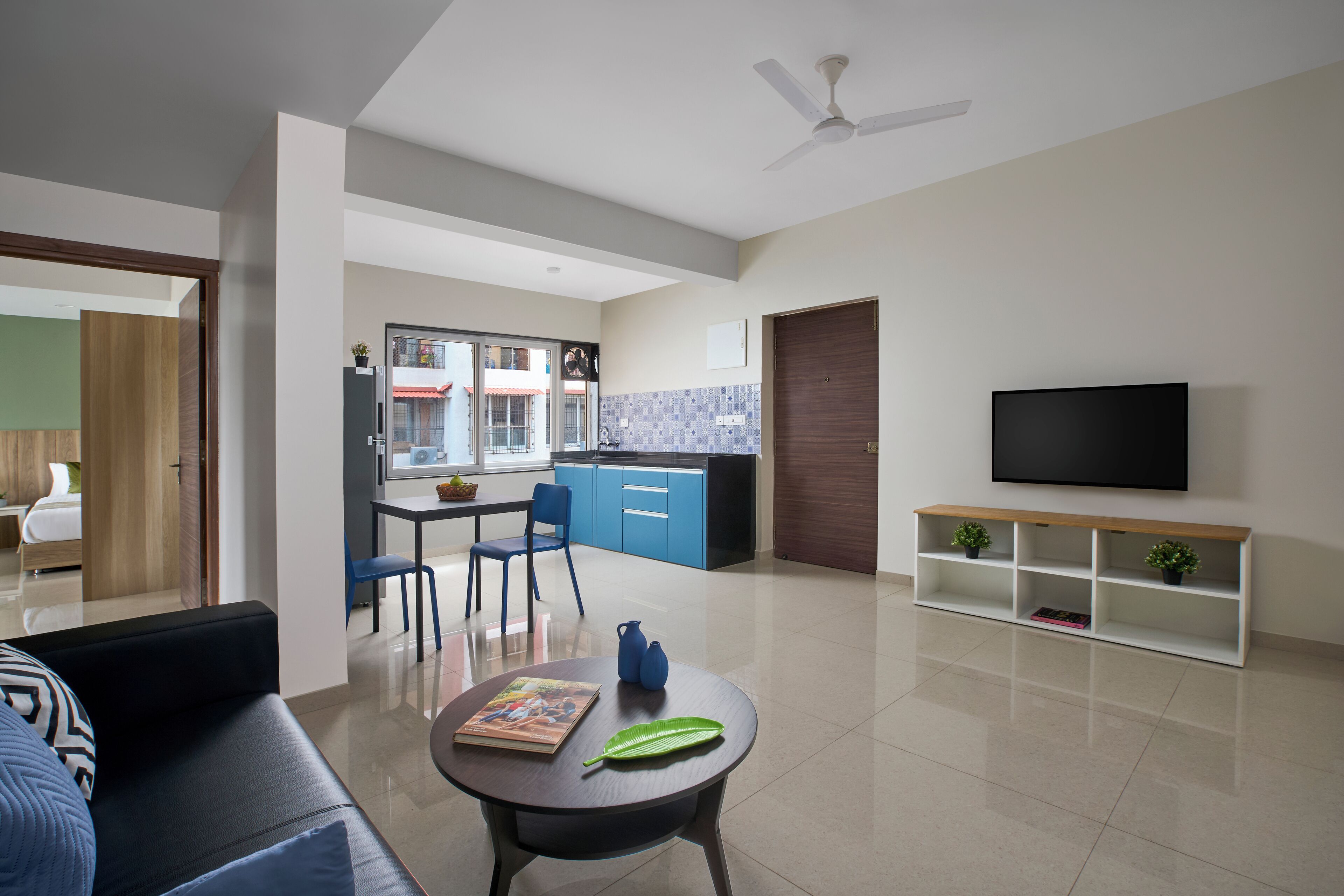 Una sala de estar moderna con un sofá, una mesa de centro con libros y una planta, un televisor sobre un mueble bajo, y una cocina al fondo.