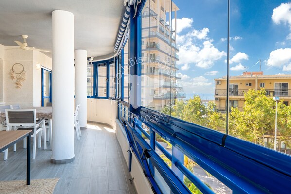 A balcony with a view of the sea, a dining area with a table and chairs, and a large window with a curtain.