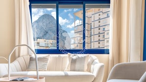 A modern living room with a sofa and armchair, a small side table, and a view of a mountain and buildings through a large window.