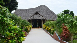 A traditional thatched-roof building with a central entrance and a pathway flanked by lush greenery. - MEREMETA LODGE (Mafia Island)