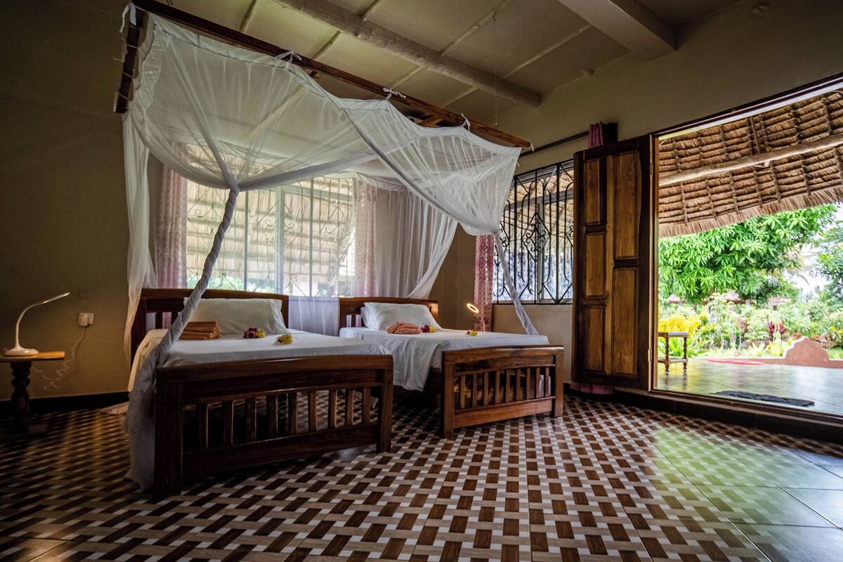 A room with two beds, a checkered floor, a window with a view of greenery, and a mosquito net over the beds.