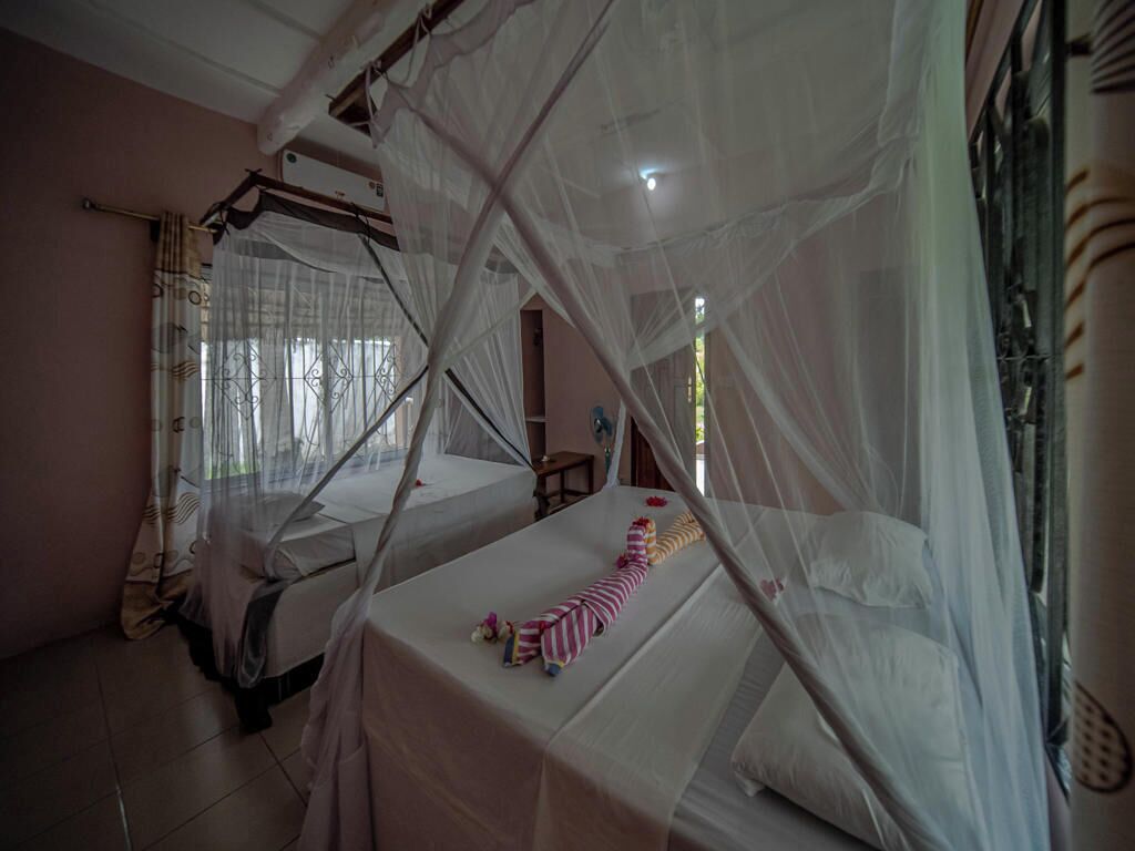 A room with two beds covered by mosquito nets, a small table with a striped cloth, and a window with curtains.