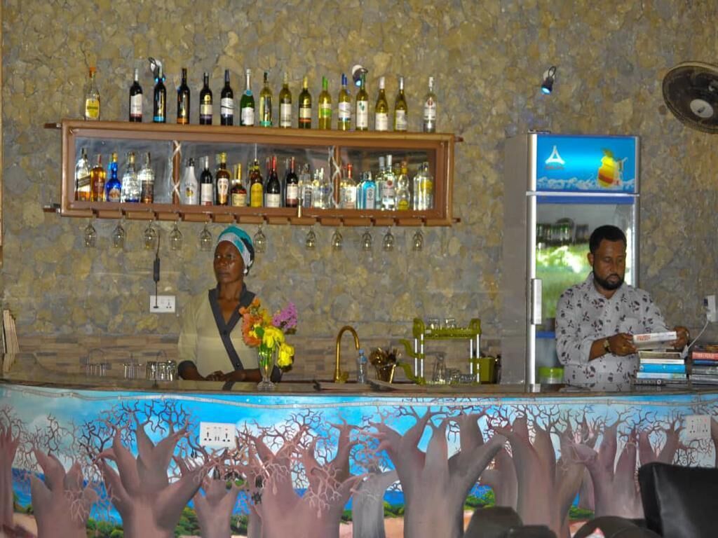 A bar area with shelves of bottles and glasses, a counter with a person, and a decorative tree mural.