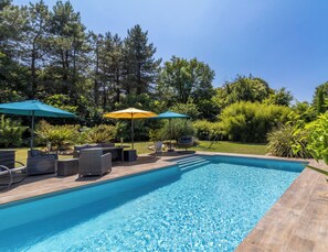 Una piscina con sillas de descanso y sombrillas, rodeada de vegetación.