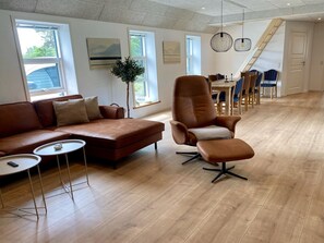 Un salon moderne avec un canapé en cuir marron, un fauteuil en cuir marron, une petite table ronde et un coin repas avec des chaises et une table en bois.