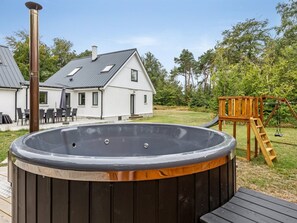 Un jacuzzi dans un jardin, avec une terrasse en bois, une maison blanche et une balançoire en bois.