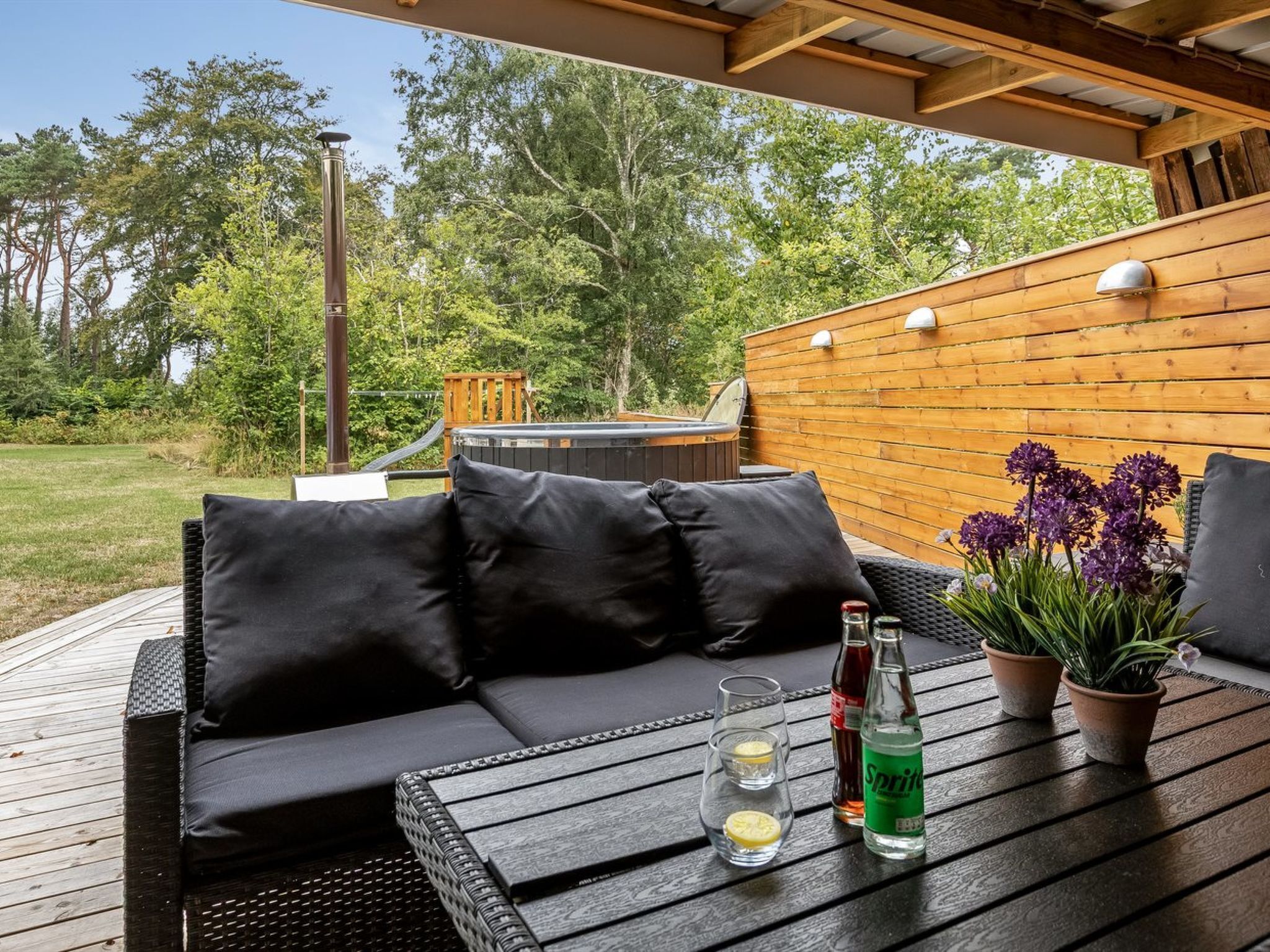 Un espace extérieur aménagé avec une terrasse en bois, un canapé noir rembourré et une table sur laquelle sont disposées des boissons et une plante en pot.