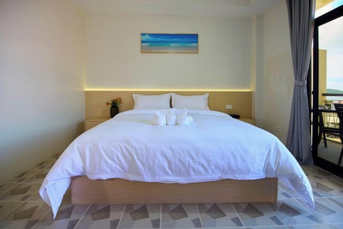 A hotel room with a large bed, a painting on the wall, and a view through the window.