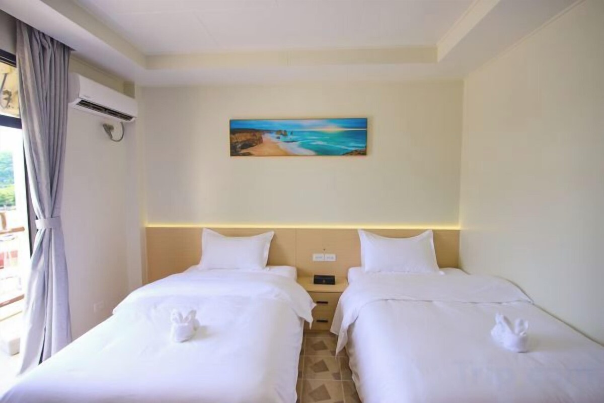 A hotel room with two beds, a painting of a beach, and a window with curtains.
