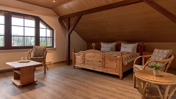 Une chambre confortable située sous les toits, avec un lit en bois, deux fauteuils en osier, une petite table et une grande fenêtre donnant sur l’extérieur.