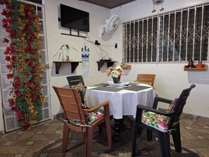 Een eethoek met een tafel gedekt voor twee personen, stoelen, een aan de muur bevestigde televisie, een decoratieve plant en een raam met decoratieve roeders.